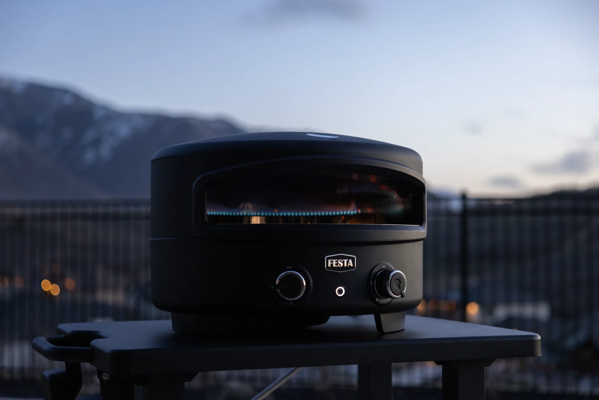 A black Festa gas pizza oven warms up on an outdoor stand during dusk with mountains in the distance.